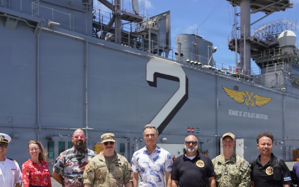 A group of nine individuals pose for a photo on the flight deck of a U.S. Navy aircraft carrier, identifiable by the large number '2' and naval aviation insignia on the ship's island structure. The group includes military personnel in various uniforms (Navy dress whites, Army/Air Force camouflage, Navy working uniform) alongside civilians in casual attire. The setting appears to be a sunny day at a naval installation.