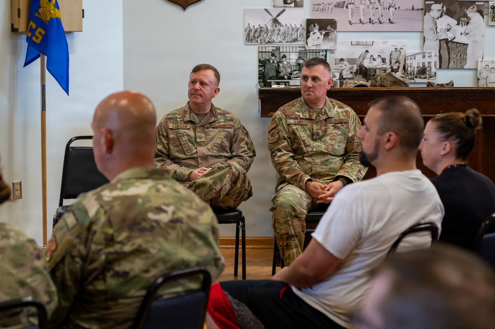 Two military personnel in OCP (Operational Camouflage Pattern) uniforms are seated at the front of a room, appearing to address or engage with an audience of mixed military and civilian attendees. The setting appears to be a briefing or informal meeting room decorated with historical black-and-white photographs on the wall. Name tapes on the uniforms read 'HANAVAN' and 'TARDY'.