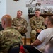Two military personnel in OCP (Operational Camouflage Pattern) uniforms are seated at the front of a room, appearing to address or engage with an audience of mixed military and civilian attendees. The setting appears to be a briefing or informal meeting room decorated with historical black-and-white photographs on the wall. Name tapes on the uniforms read 'HANAVAN' and 'TARDY'.