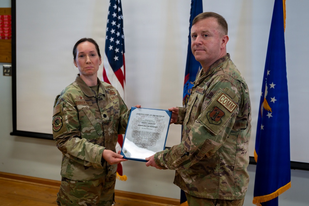 Two U.S. Air Force personnel in OCP (Operational Camouflage Pattern) uniforms are holding a Certificate of Promotion during a military promotion ceremony. The female officer on the left, identified by her name tape as 'FORSYTH' and wearing a Lieutenant Colonel oak leaf cluster, is presenting the certificate to the male recipient on the right, who wears a 'COMM' tab. The document appears to promote the recipient to Senior Master Sergeant.