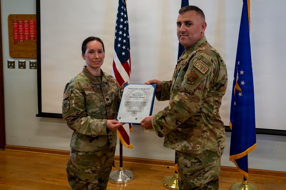 Two U.S. Air Force personnel in OCP (Operational Camouflage Pattern) uniforms are posing together while holding a Certificate of Promotion during a formal military ceremony. The female officer on the left appears to hold the rank of Lieutenant Colonel (oak leaf cluster visible), while the male on the right wears a 'COMM' unit patch. The ceremony is taking place indoors in front of an American flag and a U.S. Air Force flag.