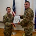 Two U.S. Air Force personnel in OCP (Operational Camouflage Pattern) uniforms are posing together while holding a Certificate of Promotion during a formal military ceremony. The female officer on the left appears to hold the rank of Lieutenant Colonel (oak leaf cluster visible), while the male on the right wears a 'COMM' unit patch. The ceremony is taking place indoors in front of an American flag and a U.S. Air Force flag.