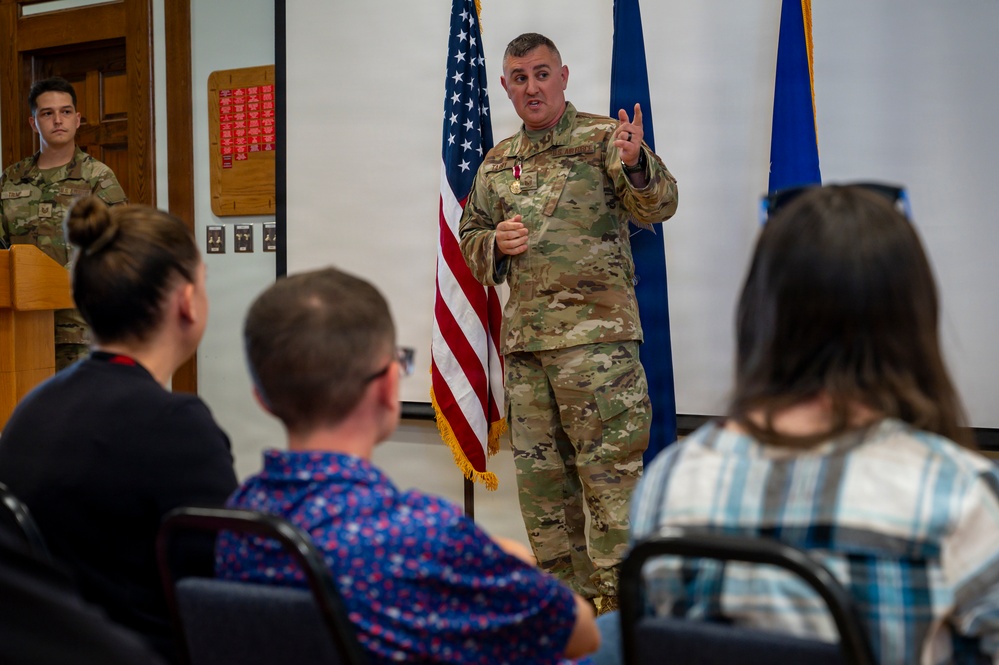A military officer in OCP (Operational Camouflage Pattern) uniform is addressing a seated audience in what appears to be a formal military ceremony or briefing room. The speaker is wearing a medal on their chest and is gesturing while speaking, with an American flag and a blue military flag visible behind them. A second uniformed service member stands at a podium in the background.