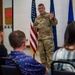 A military officer in OCP (Operational Camouflage Pattern) uniform is addressing a seated audience in what appears to be a formal military ceremony or briefing room. The speaker is wearing a medal on their chest and is gesturing while speaking, with an American flag and a blue military flag visible behind them. A second uniformed service member stands at a podium in the background.
