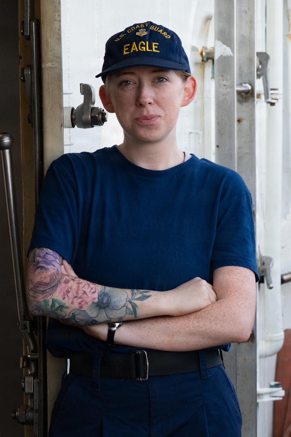 A U.S. Coast Guard service member poses with arms crossed aboard what appears to be a vessel. They are wearing a navy blue uniform and a cap embroidered with 'U.S. Coast Guard Eagle.' The subject has visible floral tattoos on their left forearm and wrist.
