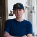 A U.S. Coast Guard service member poses with arms crossed aboard what appears to be a vessel. They are wearing a navy blue uniform and a cap embroidered with 'U.S. Coast Guard Eagle.' The subject has visible floral tattoos on their left forearm and wrist.