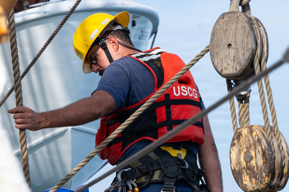 A United States Coast Guard (USCG) member is working on what appears to be a tall ship or sailing vessel, handling rigging and ropes. The individual is wearing full safety gear including a yellow hard hat, red USCG life vest, safety harness, and protective eyewear. The scene depicts active maritime maintenance or operational work near wooden block-and-tackle pulley systems.