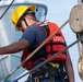 A United States Coast Guard (USCG) member is working on what appears to be a tall ship or sailing vessel, handling rigging and ropes. The individual is wearing full safety gear including a yellow hard hat, red USCG life vest, safety harness, and protective eyewear. The scene depicts active maritime maintenance or operational work near wooden block-and-tackle pulley systems.