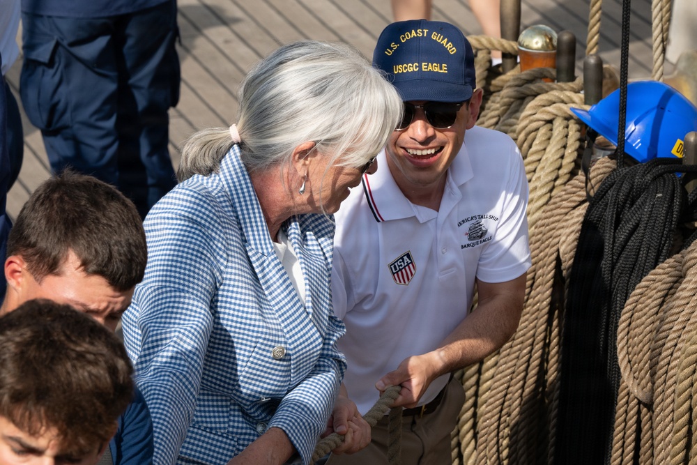 Two individuals are working together on the deck of a tall ship, handling rope rigging. The man is wearing a U.S. Coast Guard USCGC Eagle cap and a white polo shirt with 'USA' and 'America's Tallship Barque Eagle' branding. They appear to be engaged in a hands-on sailing demonstration or activity aboard the vessel.