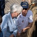 Two individuals are working together on the deck of a tall ship, handling rope rigging. The man is wearing a U.S. Coast Guard USCGC Eagle cap and a white polo shirt with 'USA' and 'America's Tallship Barque Eagle' branding. They appear to be engaged in a hands-on sailing demonstration or activity aboard the vessel.