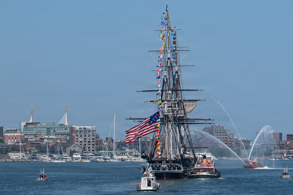 A large historic tall ship, dressed with colorful signal flags and flying a large American flag, is being escorted through a harbor by tugboats and smaller vessels. Fireboat water cannons are spraying arching jets of water on the right side, indicating a ceremonial event. A urban waterfront skyline with buildings, a marina, and what appears to be the Bunker Hill Monument is visible in the background, suggesting this is Boston Harbor.