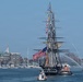 A large historic tall ship, dressed with colorful signal flags and flying a large American flag, is being escorted through a harbor by tugboats and smaller vessels. Fireboat water cannons are spraying arching jets of water on the right side, indicating a ceremonial event. A urban waterfront skyline with buildings, a marina, and what appears to be the Bunker Hill Monument is visible in the background, suggesting this is Boston Harbor.