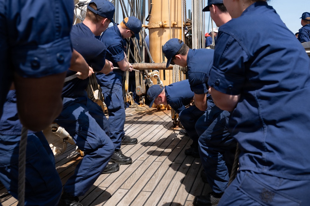 A group of personnel in matching dark navy blue uniforms are working together on the wooden deck of a sailing vessel, handling rigging and lines near a mast. The crew members appear to be engaged in coordinated physical labor, likely hauling or securing lines as part of sail handling operations. The setting is clearly aboard a tall ship or training vessel in what appears to be daylight conditions.
