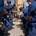 A group of personnel in matching dark navy blue uniforms are working together on the wooden deck of a sailing vessel, handling rigging and lines near a mast. The crew members appear to be engaged in coordinated physical labor, likely hauling or securing lines as part of sail handling operations. The setting is clearly aboard a tall ship or training vessel in what appears to be daylight conditions.