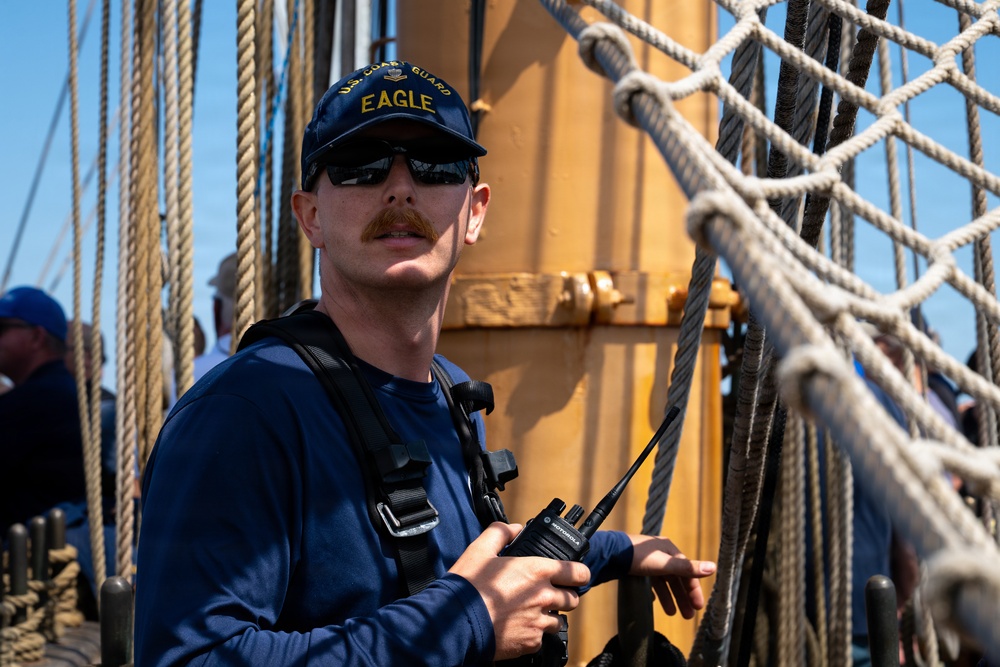 A U.S. Coast Guard crew member stands aboard the USCGC Eagle, a tall ship, holding a Motorola two-way radio. He is wearing a navy blue 'U.S. Coast Guard Eagle' cap, sunglasses, and a dark long-sleeve shirt with a radio harness. The background shows rigging, ropes, netting, and a yellow mast structure characteristic of a sailing vessel.