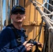 A U.S. Coast Guard crew member stands aboard the USCGC Eagle, a tall ship, holding a Motorola two-way radio. He is wearing a navy blue 'U.S. Coast Guard Eagle' cap, sunglasses, and a dark long-sleeve shirt with a radio harness. The background shows rigging, ropes, netting, and a yellow mast structure characteristic of a sailing vessel.