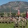 Medical Readiness Battalion welcomes new commander A U.S. Army unit conducts a formal military ceremony on a grass field, with soldiers in OCP (Operational Camouflage Pattern) uniforms rendering salutes while a color guard presents the American flag and unit guidons. The backdrop features a large rocky mountain range under a clear blue sky, consistent with the terrain near Fort Carson, Colorado. A sign in the background references an Army Community Hospital and MEDAC, suggesting a medical unit affiliation.