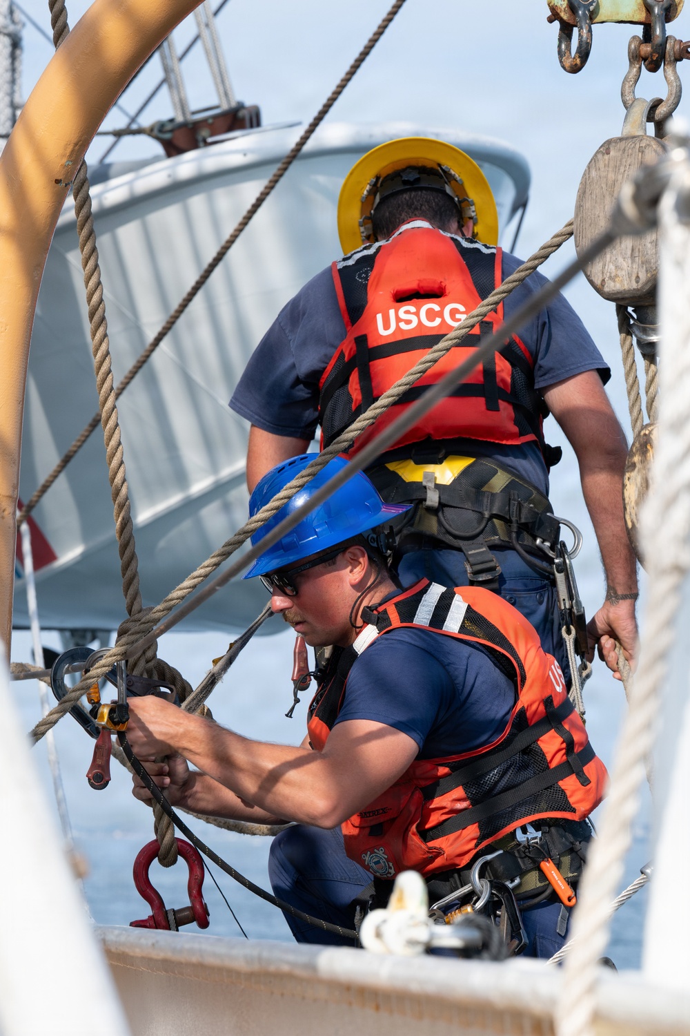 Two United States Coast Guard (USCG) personnel are working on the deck of a vessel, handling rigging and rope equipment. Both individuals are wearing orange USCG life vests, hard hats (one yellow, one blue), and safety harnesses. The foreground figure is actively working with ropes and shackles while the background figure stands facing away.