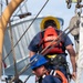 Two United States Coast Guard (USCG) personnel are working on the deck of a vessel, handling rigging and rope equipment. Both individuals are wearing orange USCG life vests, hard hats (one yellow, one blue), and safety harnesses. The foreground figure is actively working with ropes and shackles while the background figure stands facing away.