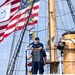 A uniformed Coast Guard officer stands with arms crossed on an elevated platform of a tall ship, surrounded by rigging and masts. Multiple flags are flying, including the United States flag and what appears to be a Coast Guard or port authority flag. The image captures a formal or ceremonial maritime setting under clear skies.