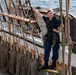 A person in navy blue uniform with a safety harness stands confidently on the rigging or rail of a tall ship, holding onto ropes overhead. The vessel appears to be underway on open water, with extensive rope work, wooden railings, and nautical equipment visible throughout. The individual appears to be a maritime professional or military/coast guard personnel based on their uniform and gear.