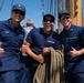 Three U.S. Coast Guard personnel pose cheerfully on the deck of a tall ship, giving thumbs up to the camera. The sailors are wearing navy blue uniforms and caps with Coast Guard insignia, with one holding a coiled rope. The setting appears to be a sunny day at a maritime event or port visit.