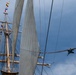 An upward-looking photograph of a tall ship's masts and rigging, decorated with colorful nautical signal flags. Several crew members are visible perched on the yardarms and rigging. A military jet aircraft is visible in the background sky to the right, suggesting this may be a ceremonial or public event.