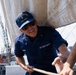 A U.S. Coast Guard cadet wearing a 'CGA 2027' cap and navy blue uniform is actively pulling a rope on what appears to be a sailing vessel. The individual is smiling and working alongside at least one other person, handling rigging or sail lines on deck. The background shows additional crew members and the open water, consistent with operations aboard a tall ship or training vessel.