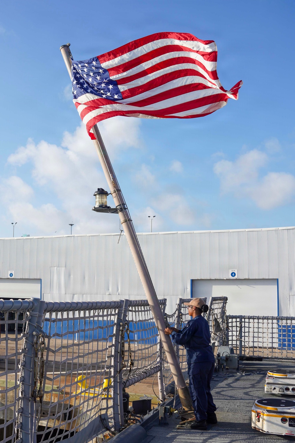 A person in a navy blue uniform is raising or handling the United States flag on what appears to be the deck of a naval or coast guard vessel docked at a port facility. The American flag is fully unfurled and waving vigorously in the wind against a partly cloudy blue sky. A large industrial warehouse or hangar building is visible in the background.