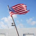 A person in a navy blue uniform is raising or handling the United States flag on what appears to be the deck of a naval or coast guard vessel docked at a port facility. The American flag is fully unfurled and waving vigorously in the wind against a partly cloudy blue sky. A large industrial warehouse or hangar building is visible in the background.