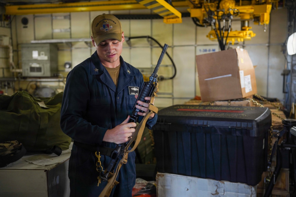 A military service member in navy blue coveralls and a tan cap is inspecting or handling a rifle with a tan sling inside what appears to be a ship's interior compartment. The individual wears rank insignia and a name tag on their coveralls. The setting appears to be a storage or working area aboard a naval vessel, with equipment, crates, and overhead crane machinery visible.