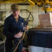 A military service member in navy blue coveralls and a tan cap is inspecting or handling a rifle with a tan sling inside what appears to be a ship's interior compartment. The individual wears rank insignia and a name tag on their coveralls. The setting appears to be a storage or working area aboard a naval vessel, with equipment, crates, and overhead crane machinery visible.