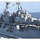 The image shows a large US Navy amphibious assault ship bearing the hull number '2', photographed at sea from a nearby vessel. The ship is underway in open ocean conditions with personnel visible along the deck railings. A red flag (appearing to be the Vietnamese national flag) is flying from one of the masts, suggesting a diplomatic or joint naval exercise context.