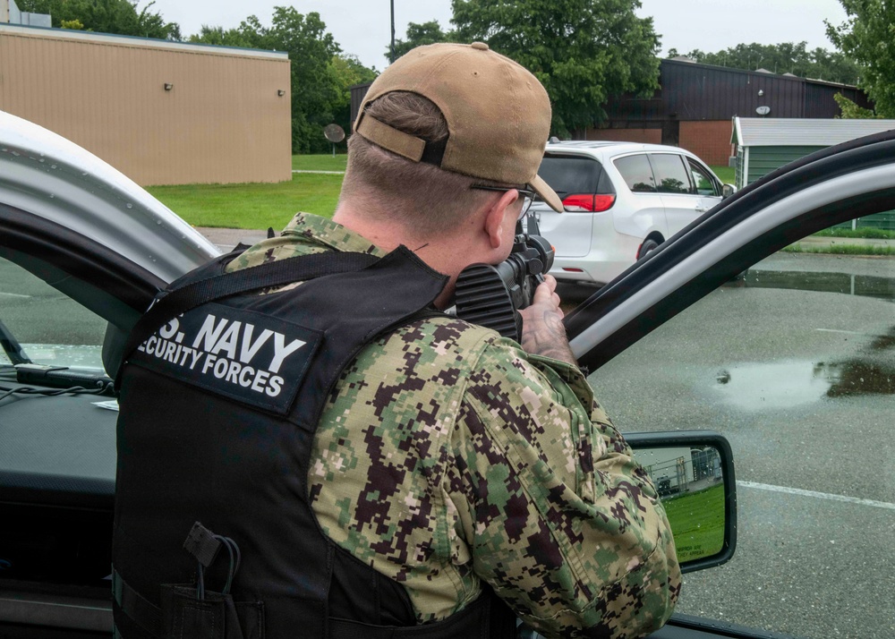 A U.S. Navy Security Forces personnel is positioned behind a vehicle door, aiming a rifle in what appears to be a training exercise or security drill. The individual is wearing Navy Working Uniform (NWU) camouflage, a tan baseball cap, sunglasses, and a black tactical vest clearly marked 'U.S. NAVY SECURITY FORCES'. The setting appears to be a military installation parking lot.