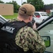 A U.S. Navy Security Forces personnel is positioned behind a vehicle door, aiming a rifle in what appears to be a training exercise or security drill. The individual is wearing Navy Working Uniform (NWU) camouflage, a tan baseball cap, sunglasses, and a black tactical vest clearly marked 'U.S. NAVY SECURITY FORCES'. The setting appears to be a military installation parking lot.