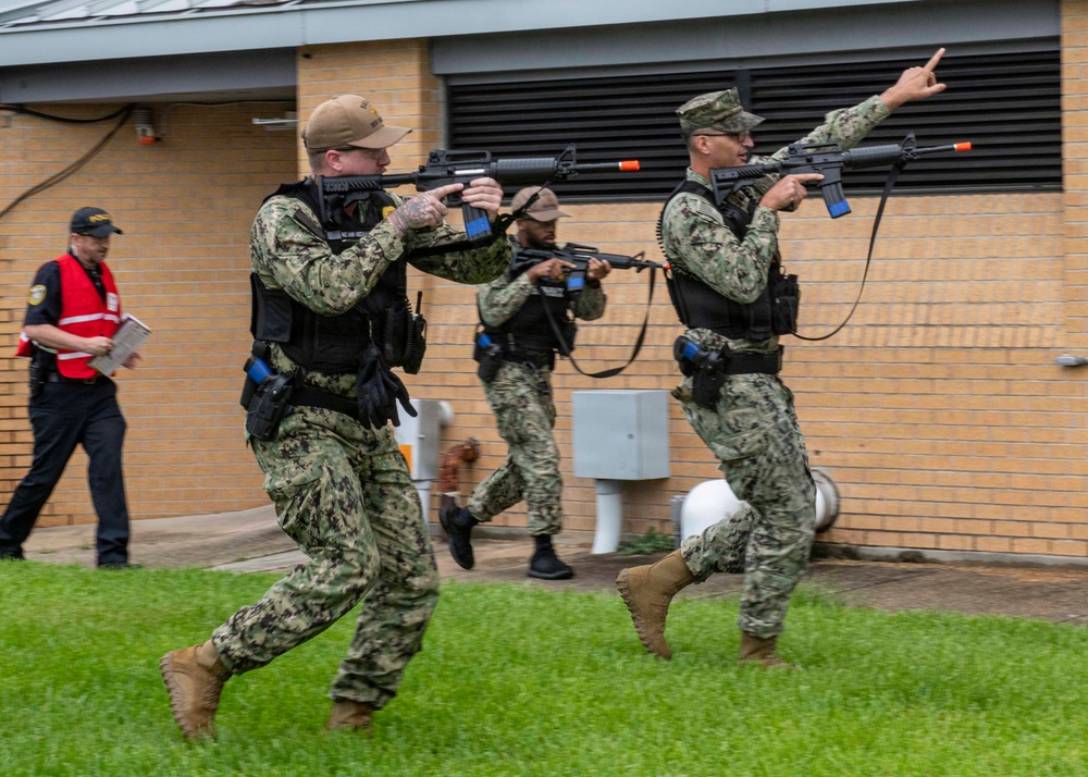 Three personnel in U.S. Navy NWU Type III (digital camouflage) uniforms are conducting what appears to be a tactical training exercise, moving in formation with rifles equipped with orange muzzle caps indicating training/simulation weapons. A civilian observer in a red vest is visible in the background taking notes, consistent with an exercise evaluator role. The orange barrel caps and blue training device attachments on the weapons confirm this is a drill rather than a live operation.