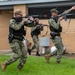 Three personnel in U.S. Navy NWU Type III (digital camouflage) uniforms are conducting what appears to be a tactical training exercise, moving in formation with rifles equipped with orange muzzle caps indicating training/simulation weapons. A civilian observer in a red vest is visible in the background taking notes, consistent with an exercise evaluator role. The orange barrel caps and blue training device attachments on the weapons confirm this is a drill rather than a live operation.
