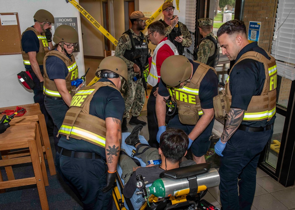 The image depicts what appears to be a mass casualty training exercise or drill involving fire department personnel and U.S. Navy military members inside a building. Firefighters wearing tactical vests and ballistic helmets are attending to a simulated victim on a stretcher, while Navy personnel and a safety observer in a red vest look on. Yellow police tape is visible in the background, indicating a controlled exercise perimeter.