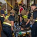 The image depicts what appears to be a mass casualty training exercise or drill involving fire department personnel and U.S. Navy military members inside a building. Firefighters wearing tactical vests and ballistic helmets are attending to a simulated victim on a stretcher, while Navy personnel and a safety observer in a red vest look on. Yellow police tape is visible in the background, indicating a controlled exercise perimeter.