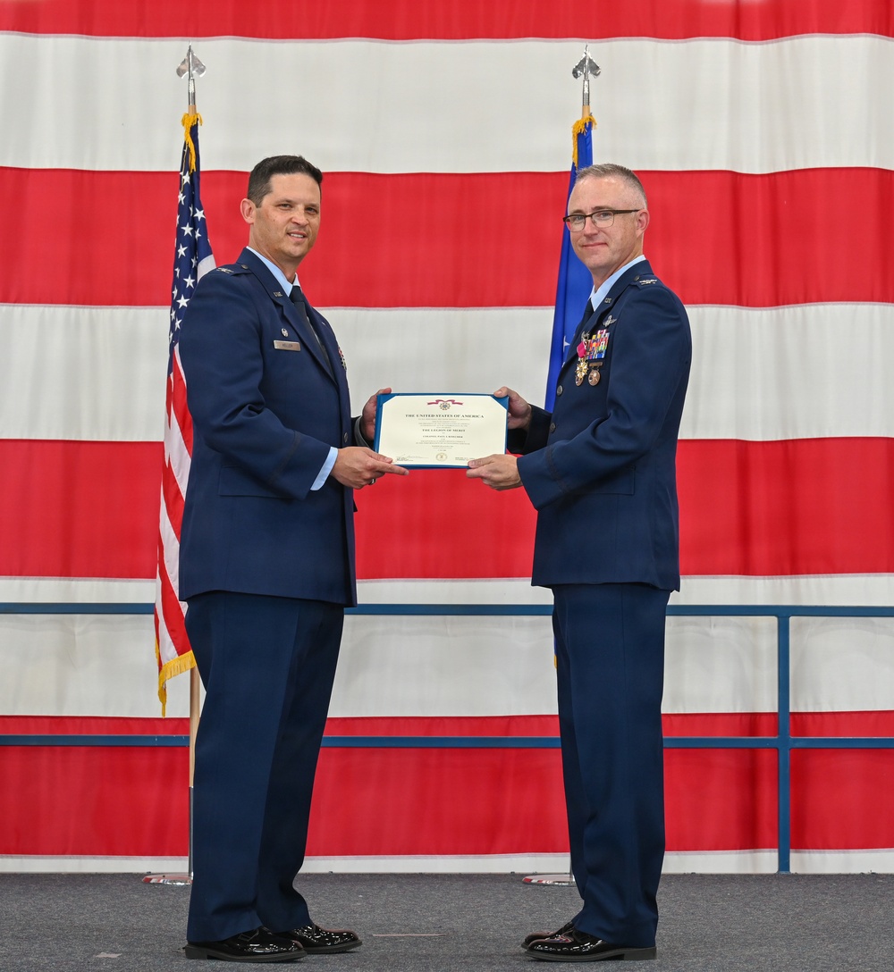 Two U.S. Air Force officers in dress blue uniforms are jointly holding a certificate during a formal military ceremony. The certificate appears to be a Legion of Merit award, as indicated by the visible text on the document. The ceremony is taking place in front of a large American flag backdrop alongside an Air Force flag.