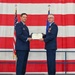 Two U.S. Air Force officers in dress blue uniforms are jointly holding a certificate during a formal military ceremony. The certificate appears to be a Legion of Merit award, as indicated by the visible text on the document. The ceremony is taking place in front of a large American flag backdrop alongside an Air Force flag.