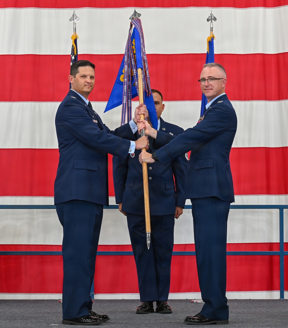A military change of command ceremony is taking place in what appears to be an aircraft hangar or large military facility. Two U.S. Air Force officers in service dress uniforms are jointly holding a unit guidon (flag on a staff), which is the central symbolic act of a change of command. A large American flag serves as the backdrop, and a third uniformed airman stands between them.
