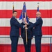 A military change of command ceremony is taking place in what appears to be an aircraft hangar or large military facility. Two U.S. Air Force officers in service dress uniforms are jointly holding a unit guidon (flag on a staff), which is the central symbolic act of a change of command. A large American flag serves as the backdrop, and a third uniformed airman stands between them.
