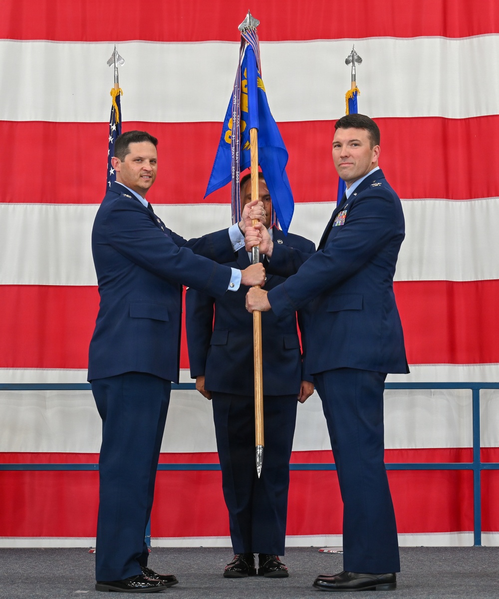 A military change of command ceremony is taking place, with two U.S. Air Force officers jointly holding a unit guidon (flag on a staff) in a formal transfer of command. The ceremony is conducted in front of a large American flag backdrop, with a third uniformed individual visible in the background. This ritual symbolizes the transfer of authority and responsibility from one commanding officer to another.
