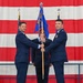A military change of command ceremony is taking place, with two U.S. Air Force officers jointly holding a unit guidon (flag on a staff) in a formal transfer of command. The ceremony is conducted in front of a large American flag backdrop, with a third uniformed individual visible in the background. This ritual symbolizes the transfer of authority and responsibility from one commanding officer to another.