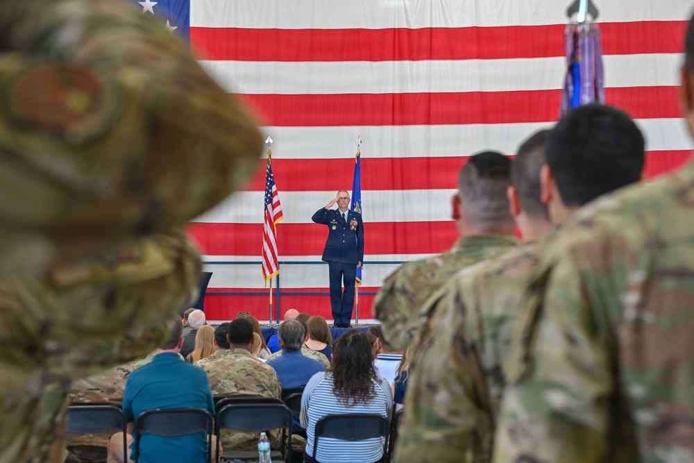 A military ceremony is taking place inside what appears to be a hangar or large indoor facility. A uniformed officer in blue dress uniform stands at the front saluting, flanked by American and Air Force flags against a large American flag backdrop. An audience of military personnel in camouflage uniforms and civilians are seated and standing in attendance.