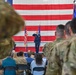 A military ceremony is taking place inside what appears to be a hangar or large indoor facility. A uniformed officer in blue dress uniform stands at the front saluting, flanked by American and Air Force flags against a large American flag backdrop. An audience of military personnel in camouflage uniforms and civilians are seated and standing in attendance.
