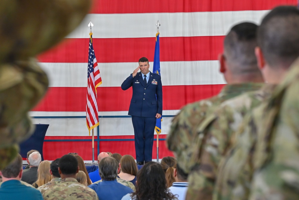 A U.S. Air Force officer in dress blues is rendering a salute on a stage during what appears to be a formal military ceremony, likely a change of command or activation ceremony. The event is held in a hangar or large indoor venue decorated with a large American flag backdrop. An audience of both uniformed military personnel and civilians is present.