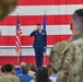 A U.S. Air Force officer in dress blues is rendering a salute on a stage during what appears to be a formal military ceremony, likely a change of command or activation ceremony. The event is held in a hangar or large indoor venue decorated with a large American flag backdrop. An audience of both uniformed military personnel and civilians is present.