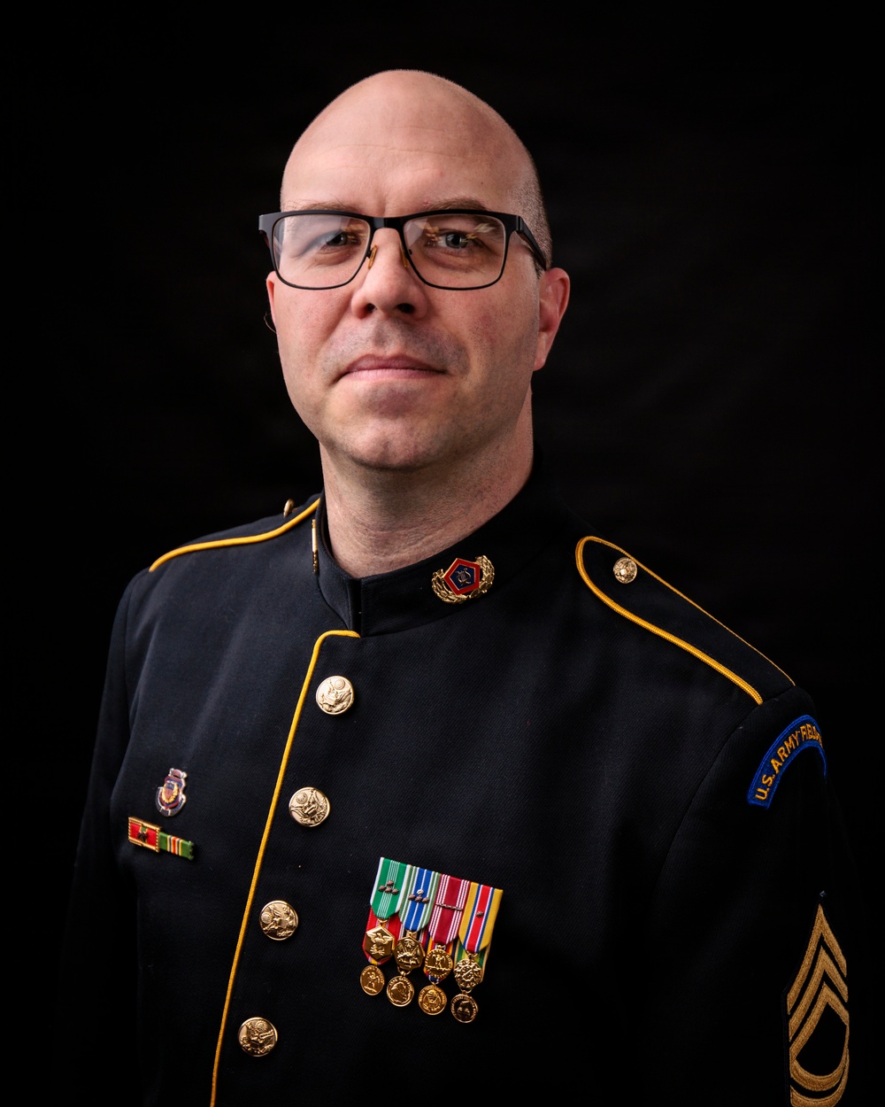 A bald male soldier wearing a U.S. Army dress uniform (black with gold piping) poses for a formal portrait against a black background. He wears glasses and displays multiple medals, ribbons, and insignia consistent with a senior non-commissioned officer rank. The uniform includes a sleeve patch reading 'U.S. Army' and sergeant first class or master sergeant chevrons on the sleeve.